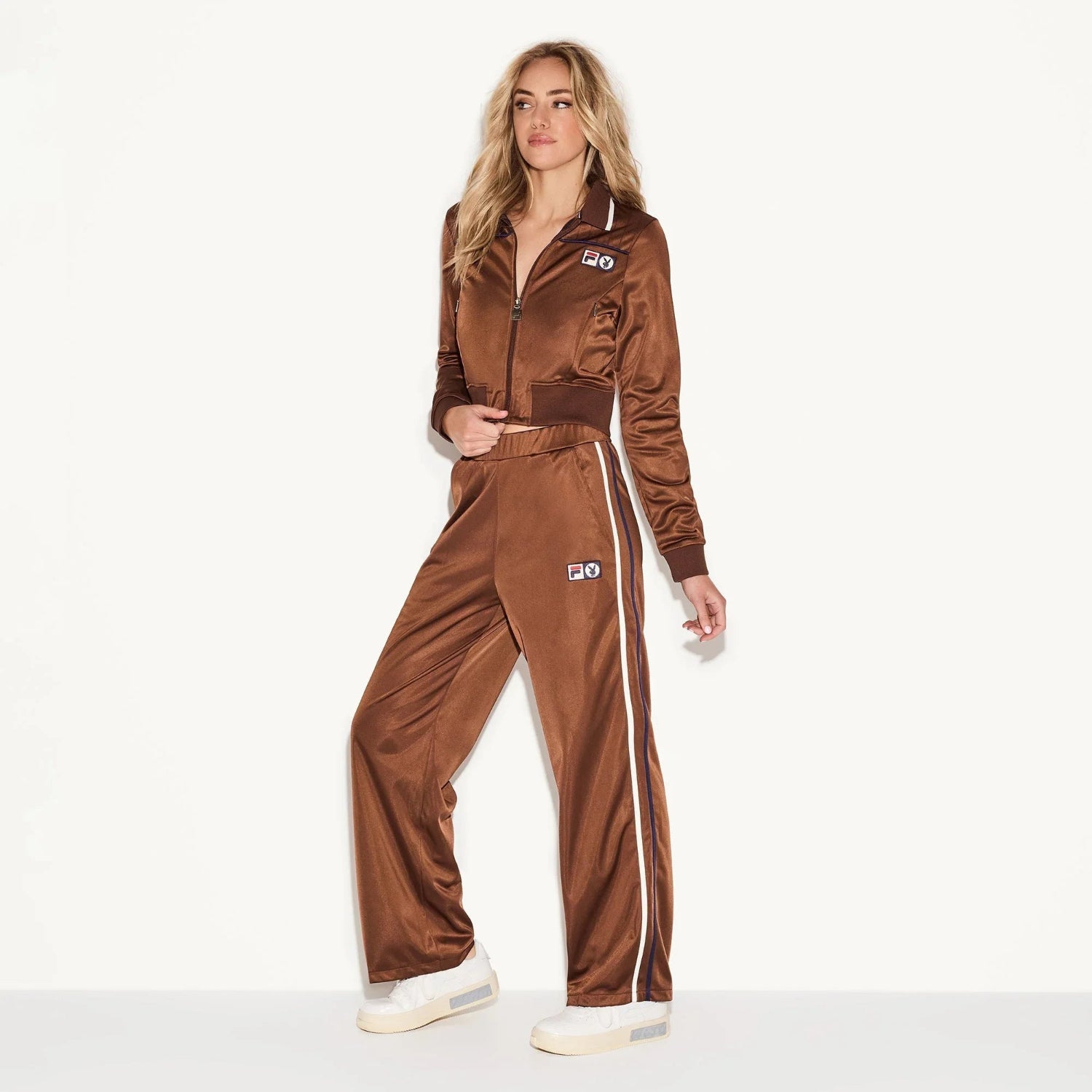 Woman wearing brown track jacket and pants set with white side stripes and logo patches