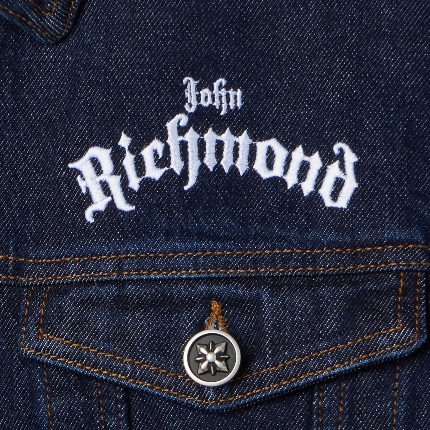Close-up of blue denim jacket pocket with white John Richmond embroidery and metal button