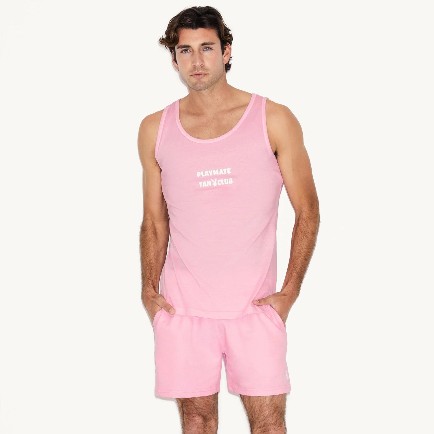 Man wearing pink sweat shorts and tank top with 'Playmate Fan Club' graphic, hands in pockets
