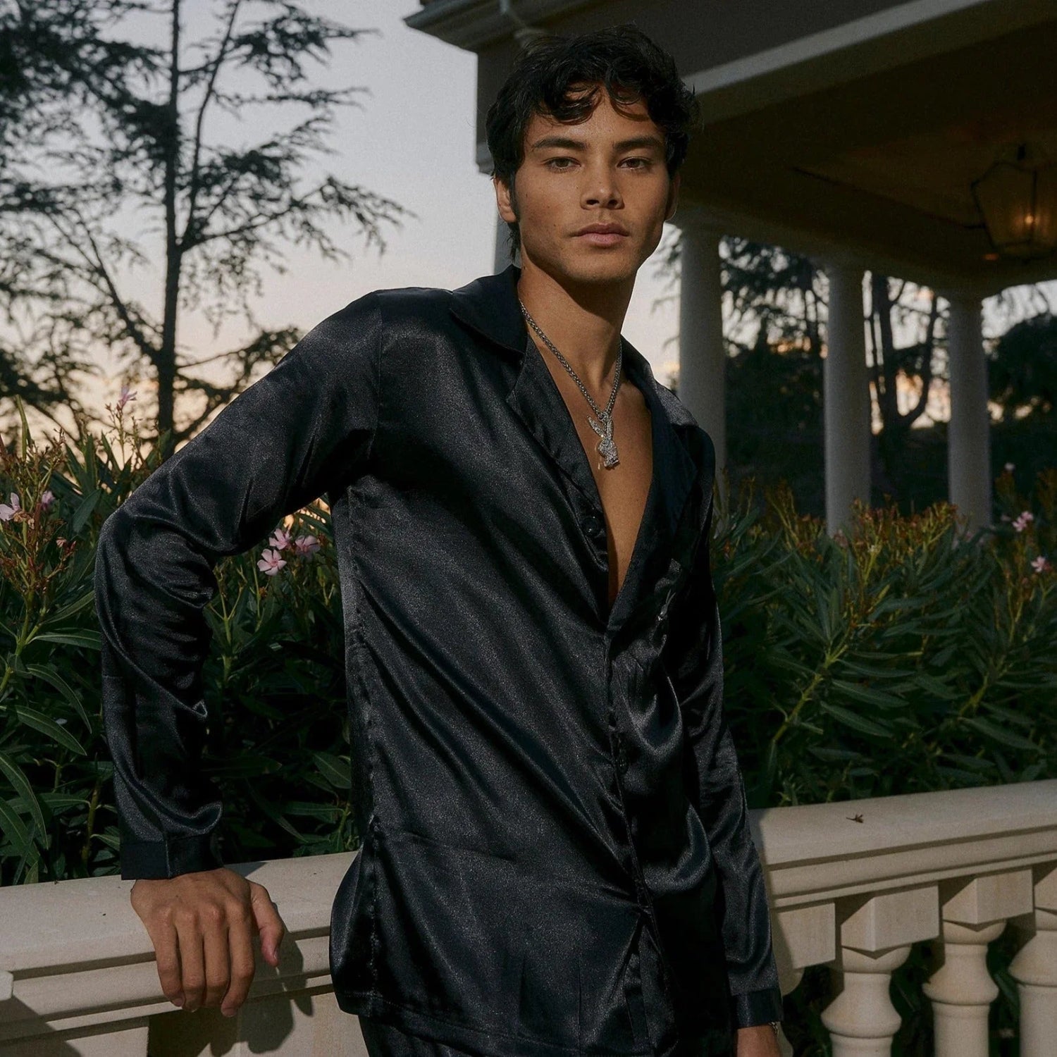 Man wearing black satin lounge set standing outdoors by a mansion railing at dusk.