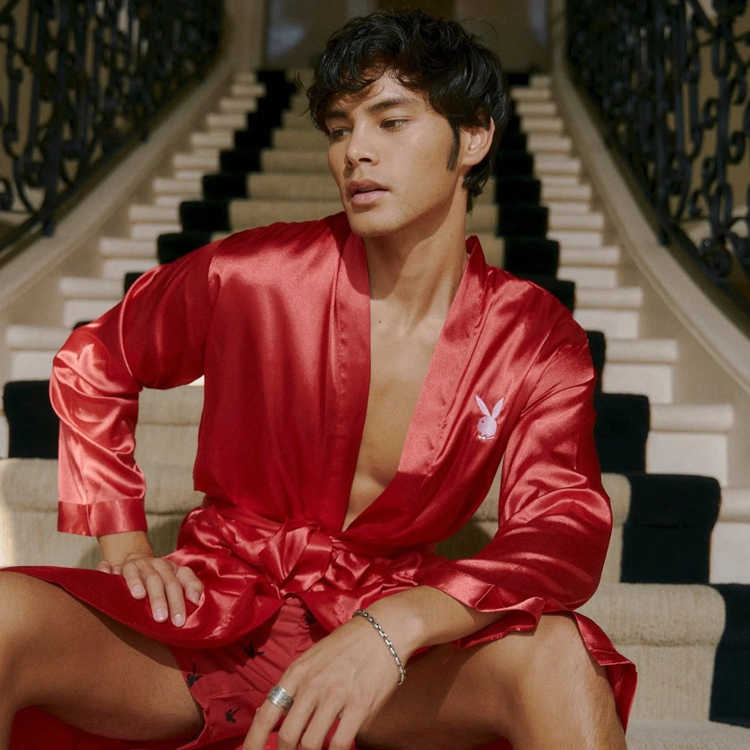 Man wearing a red satin robe with logo, seated on a staircase indoors