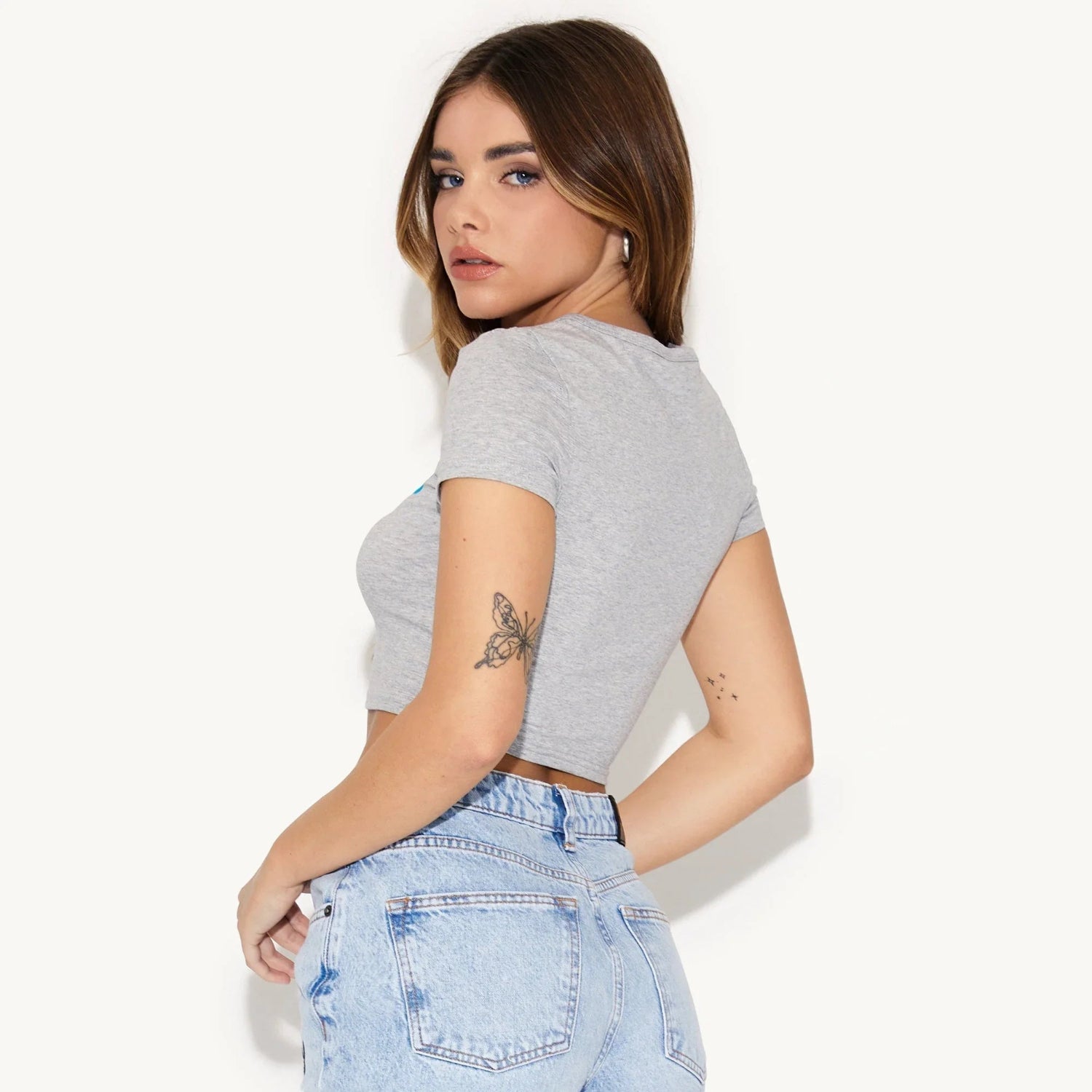 Woman wearing a grey crop baby tee and high-waisted light wash jeans, showing butterfly tattoo.