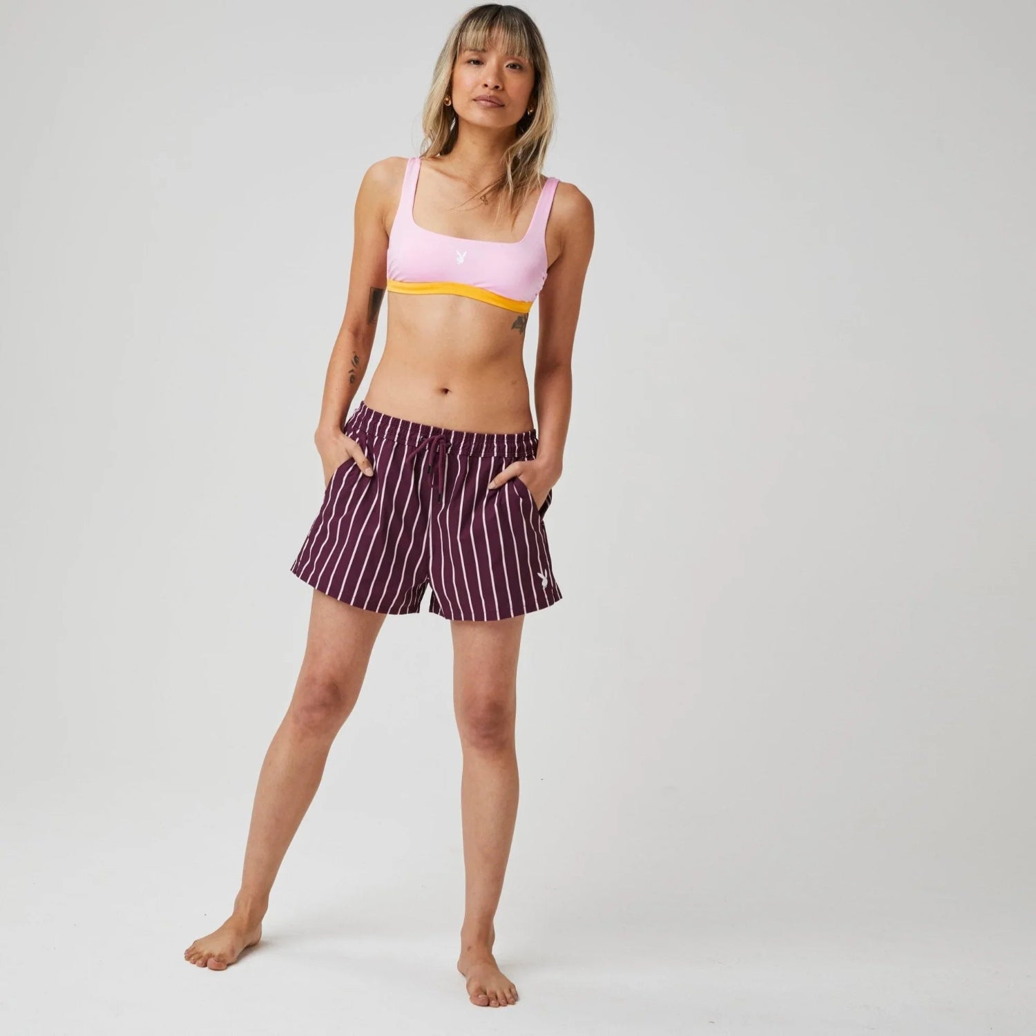 Woman wearing pink bikini top and maroon striped swim trunks with pockets, standing barefoot