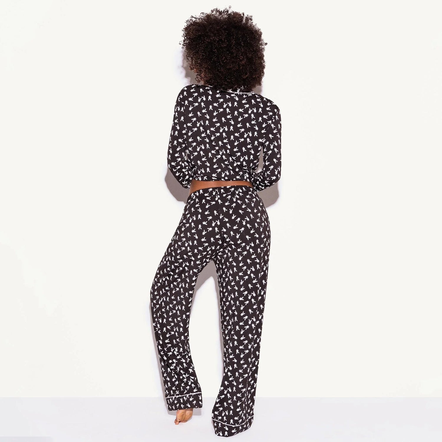 Woman wearing black pajama set with white all-over print, facing away, on white background.