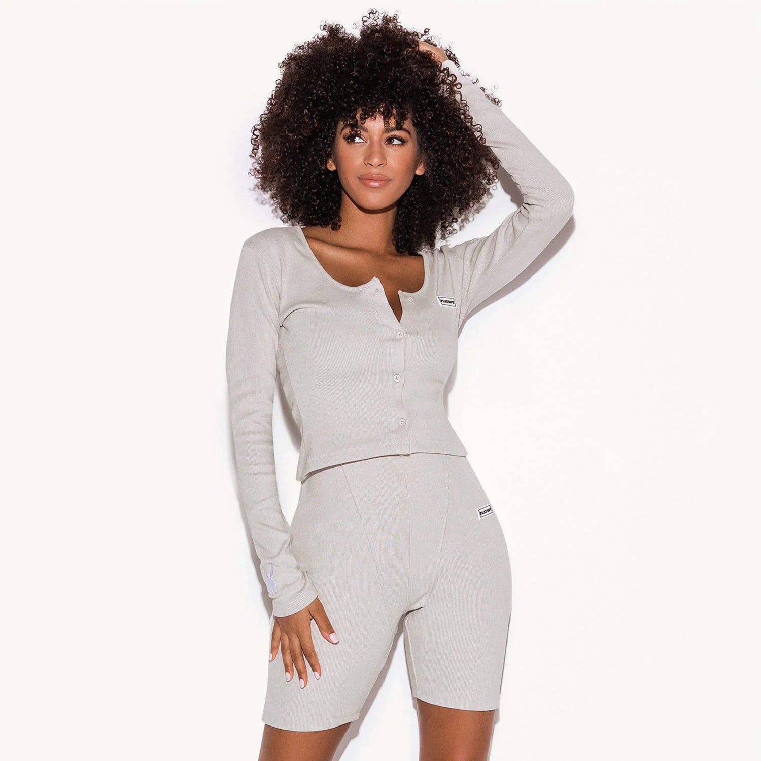 Woman wearing light grey ribbed lounge set with long sleeve top and bike shorts against white background