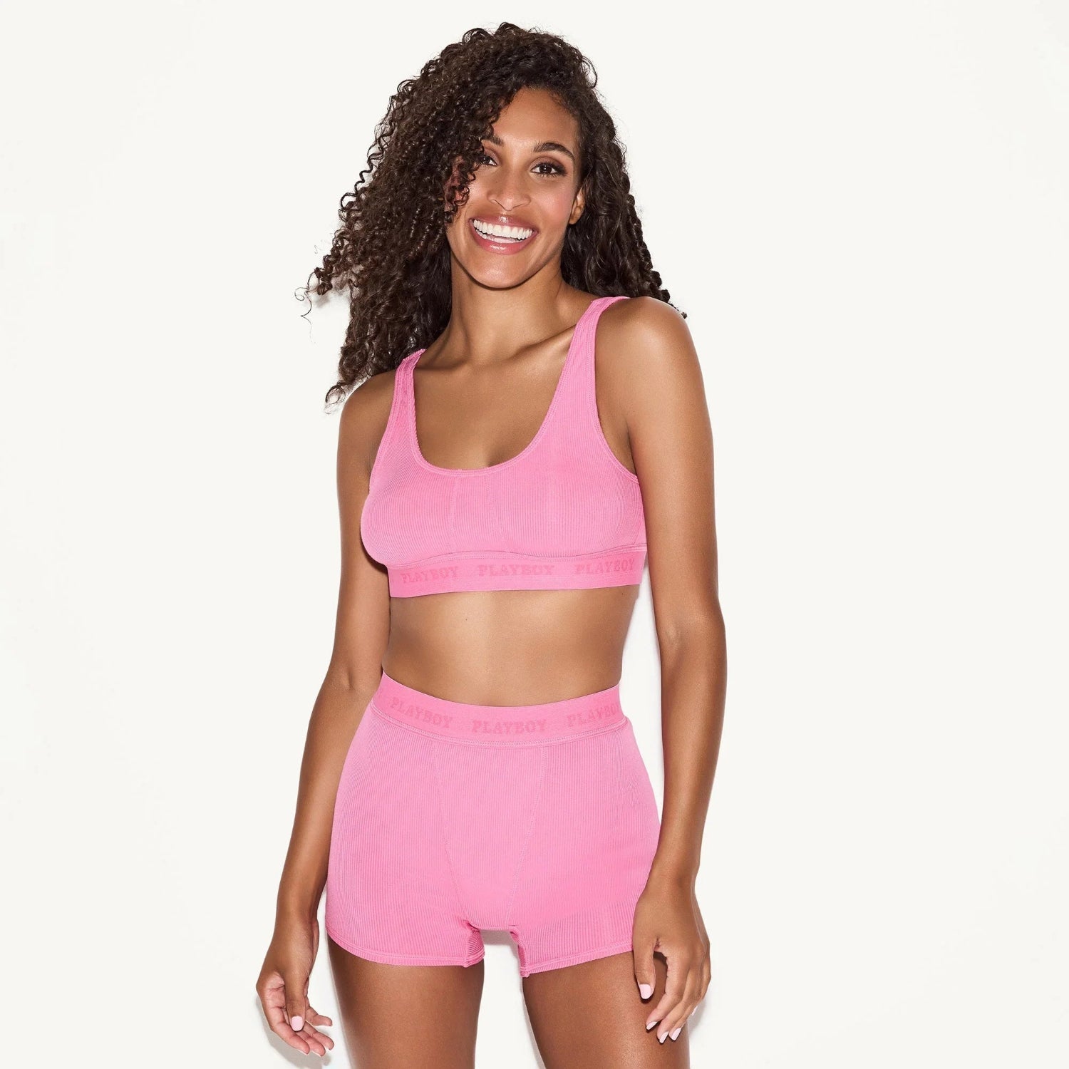Woman wearing pink ribbed sports bra and shorts set with logo waistband, standing and smiling