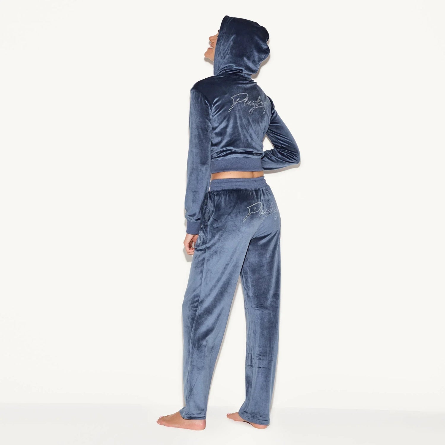 Woman wearing blue velour tracksuit with hood, embroidery on back and pants, standing barefoot