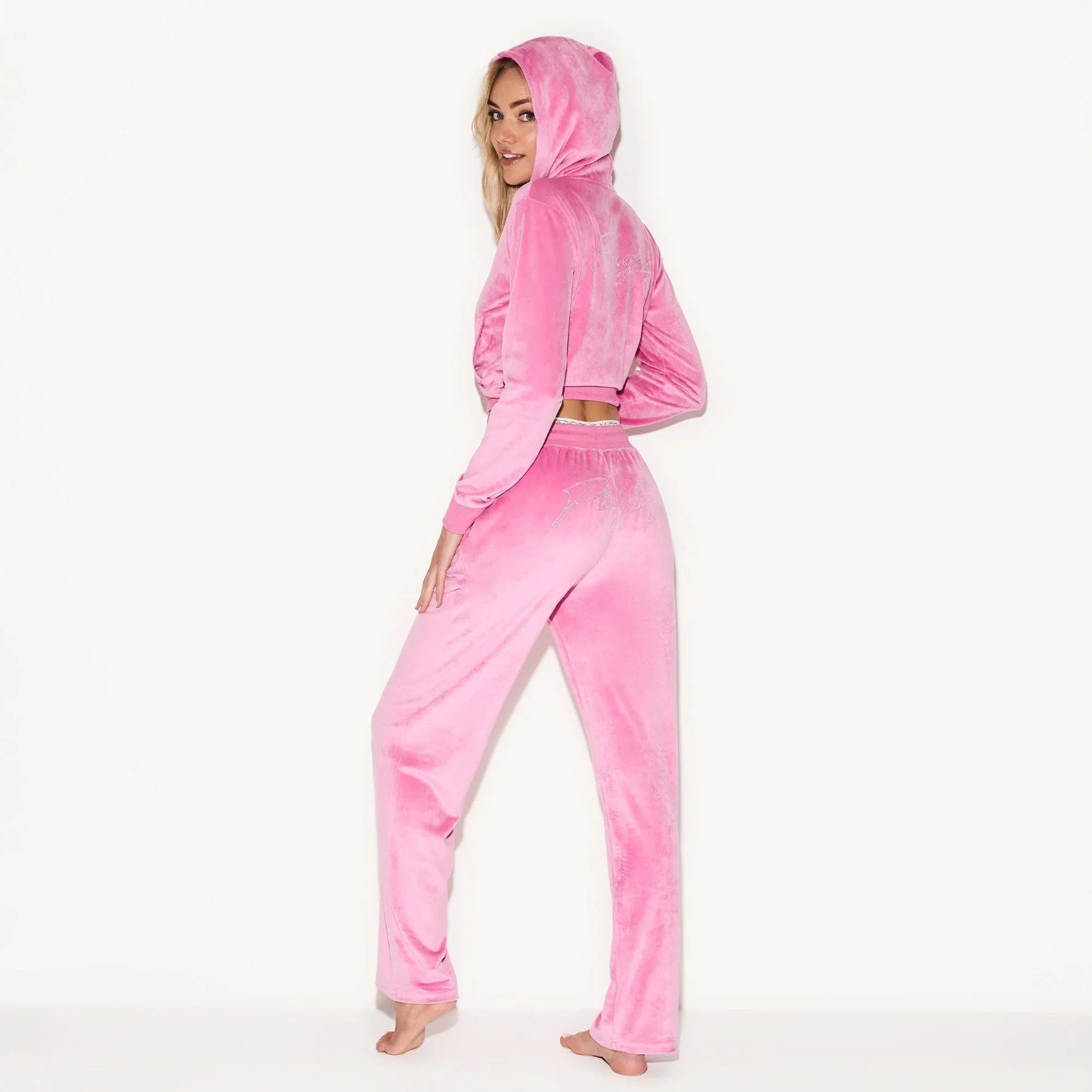 Woman wearing pink velour tracksuit with hood, long sleeves, and full-length pants, posing barefoot