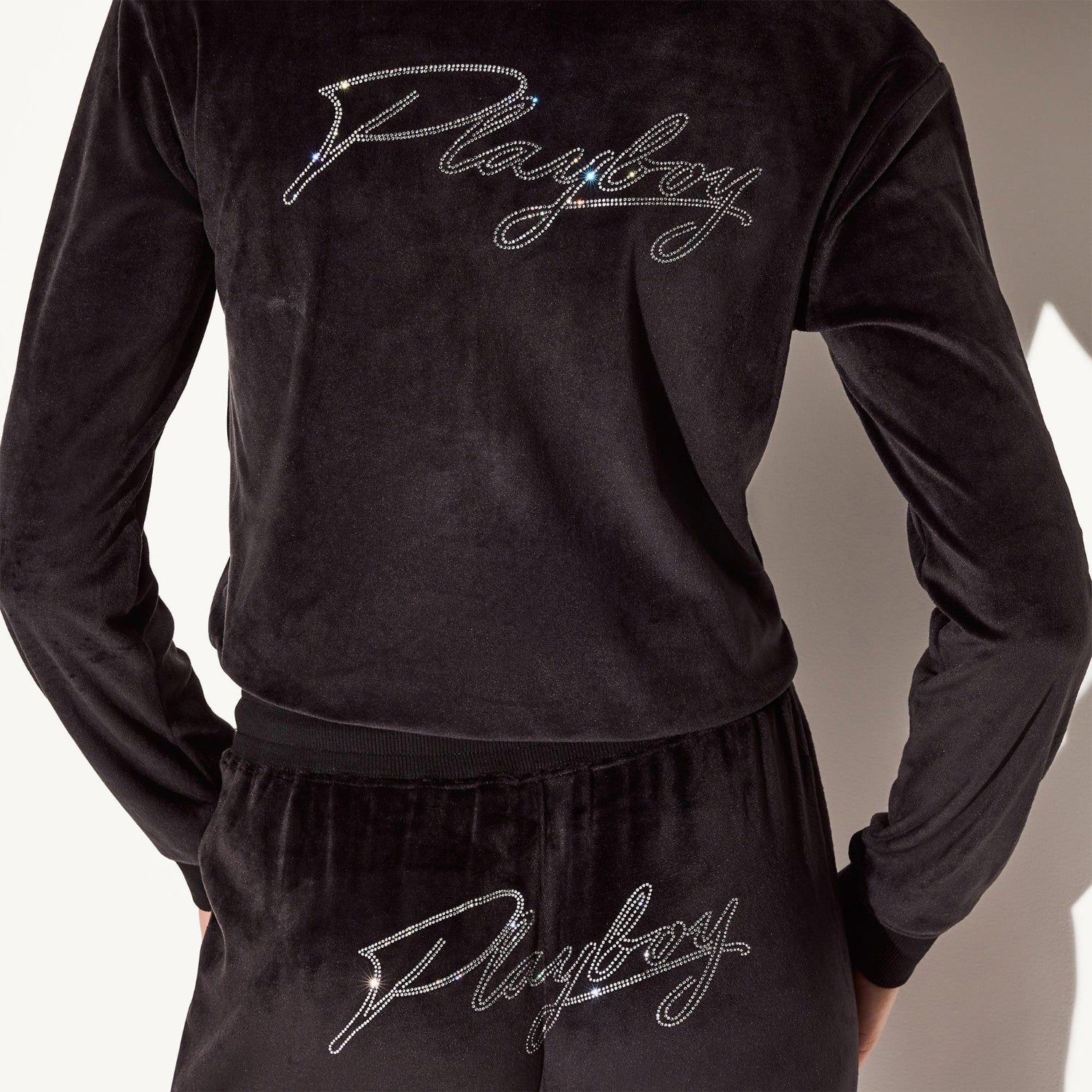 Woman wearing black velour pants and jacket with rhinestone script logo on back and waistband