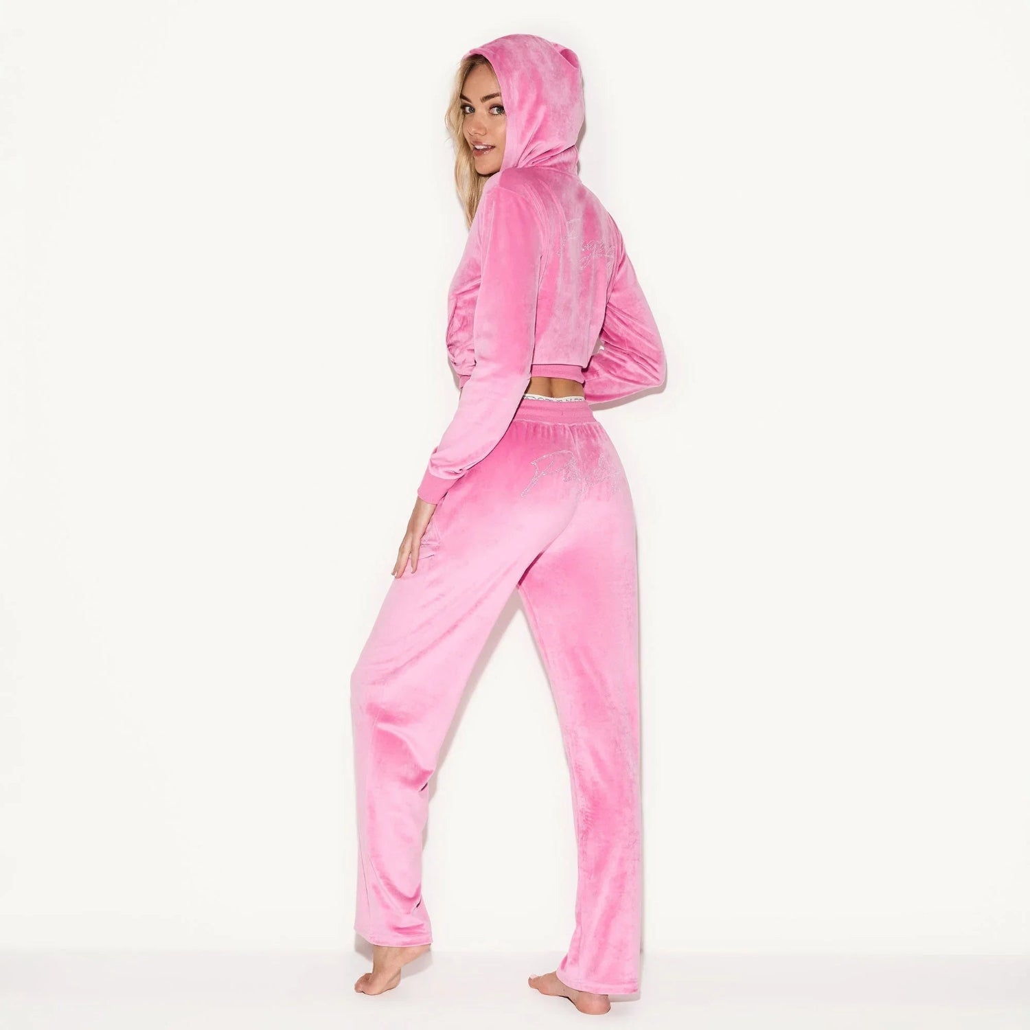 Woman posing in pink velour pants and matching hoodie, standing barefoot against white background